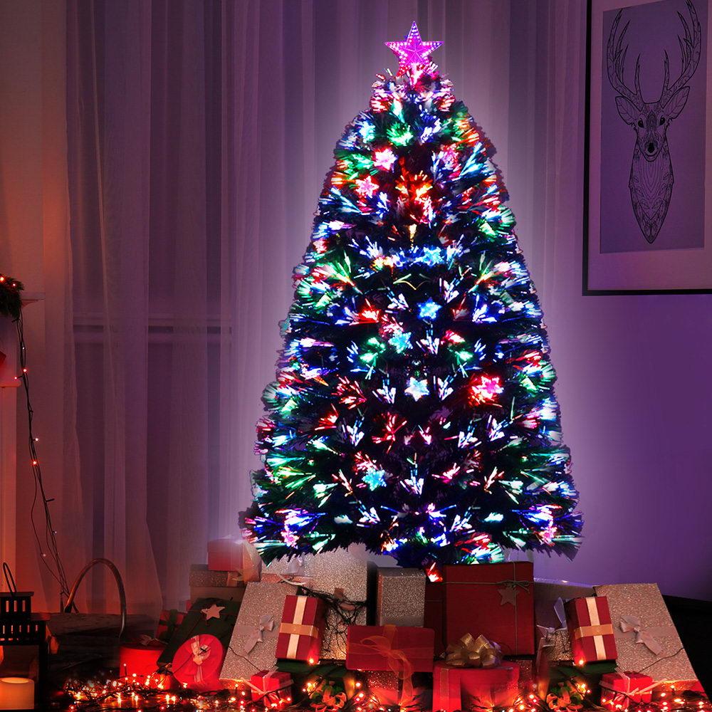 A decorated Christmas tree stands in a living room setting, illuminated with multicolored star-shaped lights and surrounded by wrapped gifts at the base.