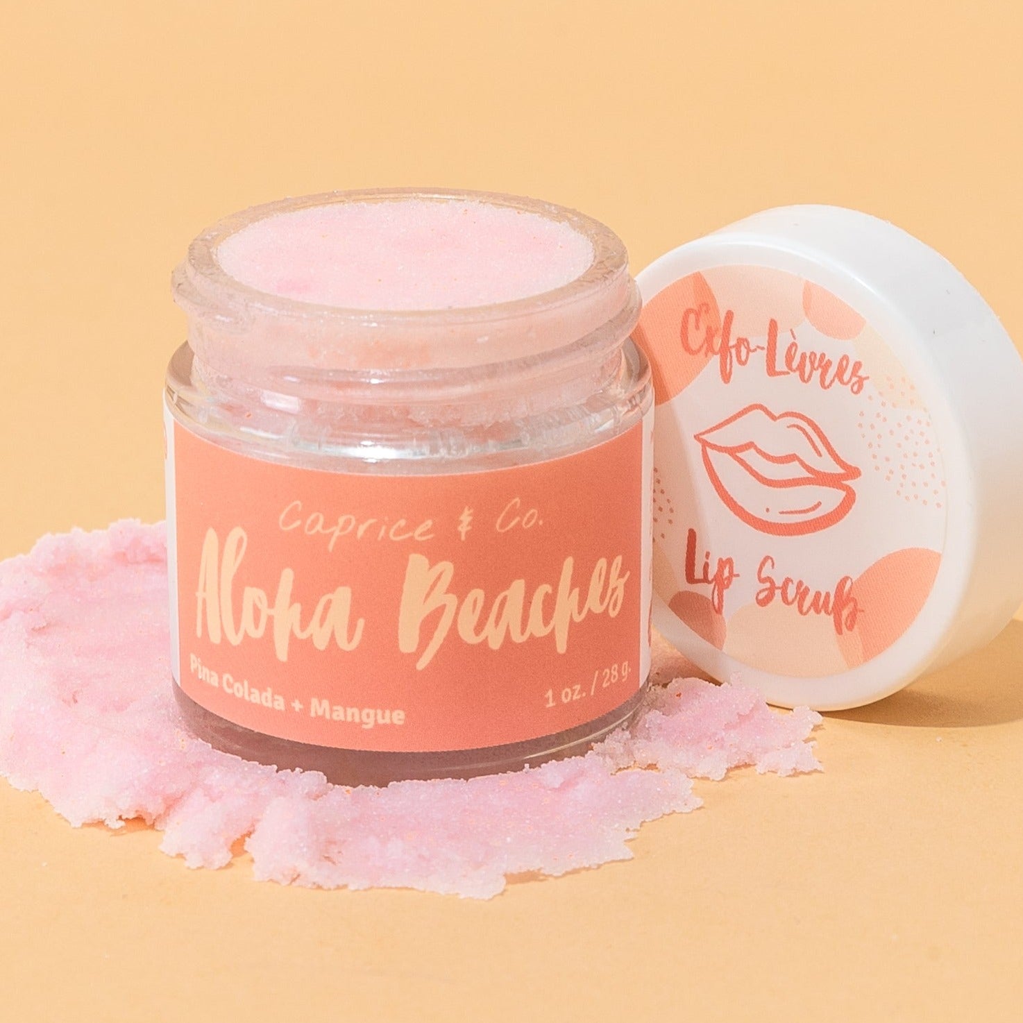 A jar of Caprice & Co. Aloha Beaches lip scrub with pink scrub visible inside, shown next to its white lid on a warm peach background with scattered pink scrub. Best online retail store. Axis-Dynamics
