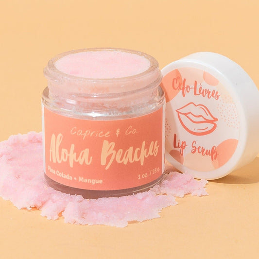 A jar of Caprice & Co. Aloha Beaches lip scrub with pink scrub visible inside, shown next to its white lid on a warm peach background with scattered pink scrub. Best online retail store. Axis-Dynamics