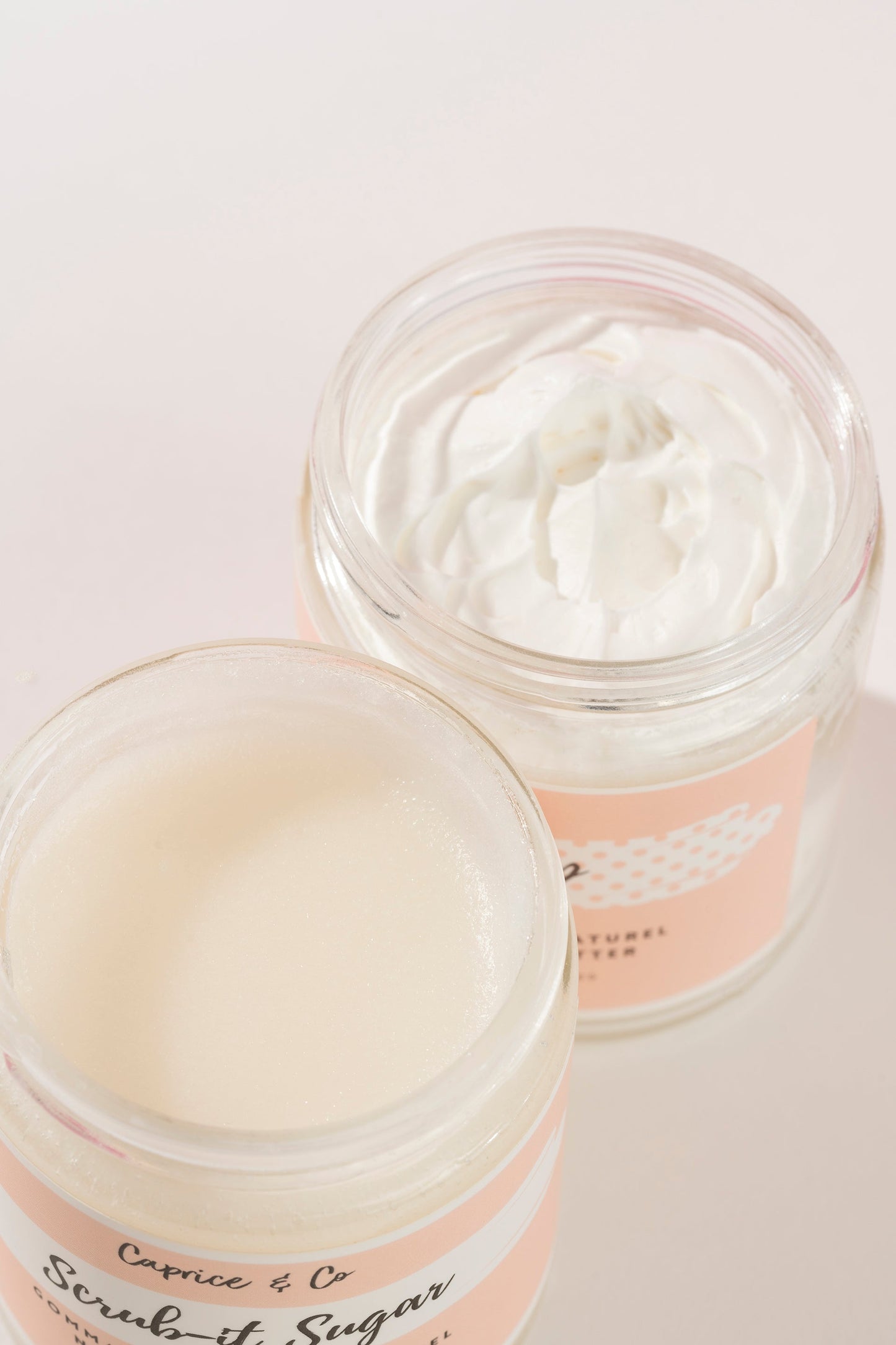 Two open glass jars of Caprice & Co. Scrub-it Sugar body scrub, showing the thick white cream product inside, set against a clean, light pink background. fragrance free body lotion for eczema relief. Online retail shops. Axis-Dynamics