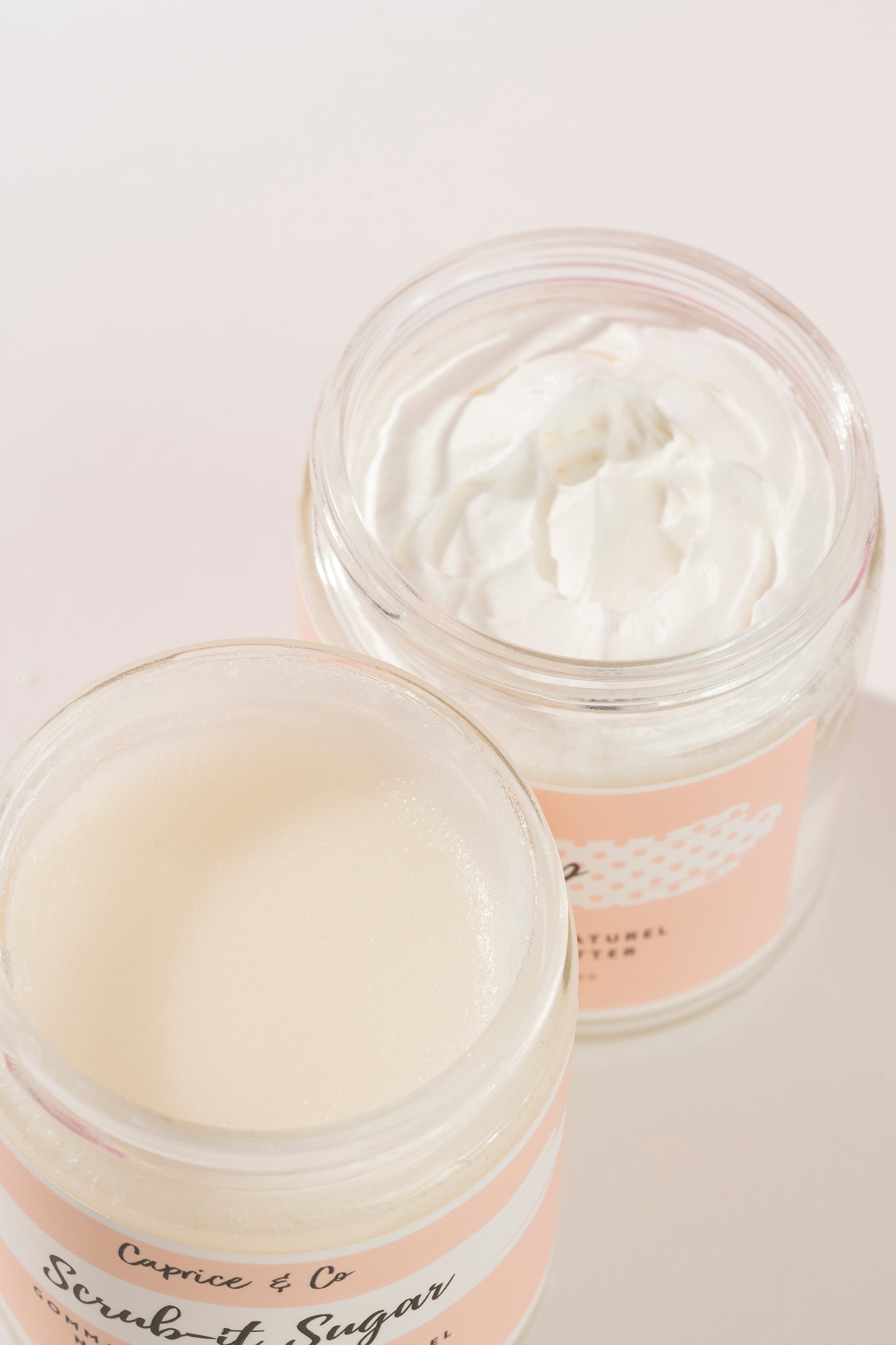 Two open glass jars of Caprice & Co. Scrub-it Sugar body scrub, showing the thick white cream product inside, set against a clean, light pink background. fragrance free body lotion for eczema relief. Online retail shops. Axis-Dynamics