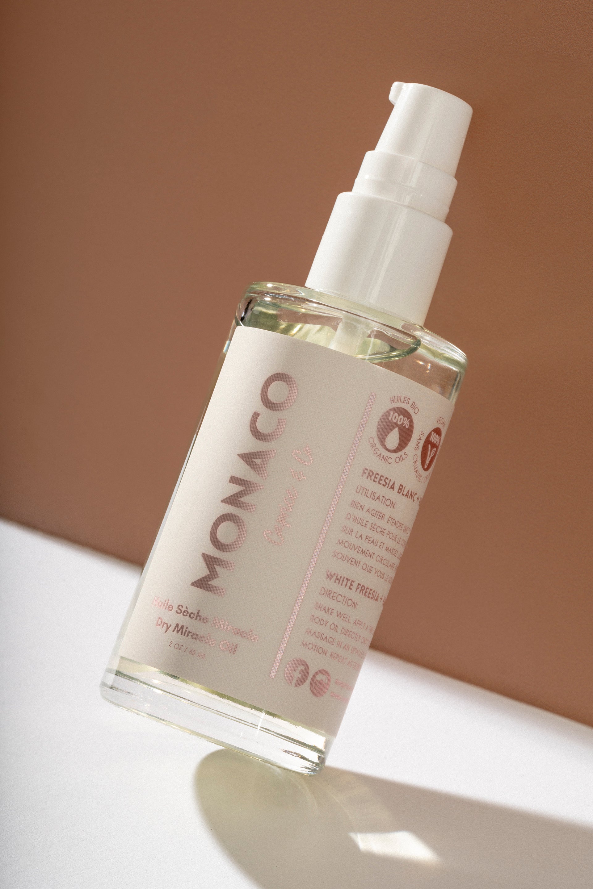 A clear pump bottle of Monaco Freesia Hair Oil with a white label and white pump dispenser, set against a warm brown background on a white surface. deep hydrating body butter for winter dry skin