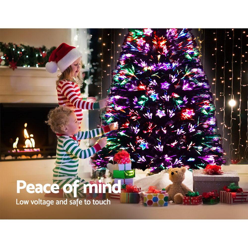 A festive Christmas tree lit with multicolored lights, with two children in holiday clothing standing beside it in a decorated living room. Presents, a teddy bear, and warm string lights are visible, with the text 'Peace of mind Low voltage and safe to touch' overlaid at the bottom left.