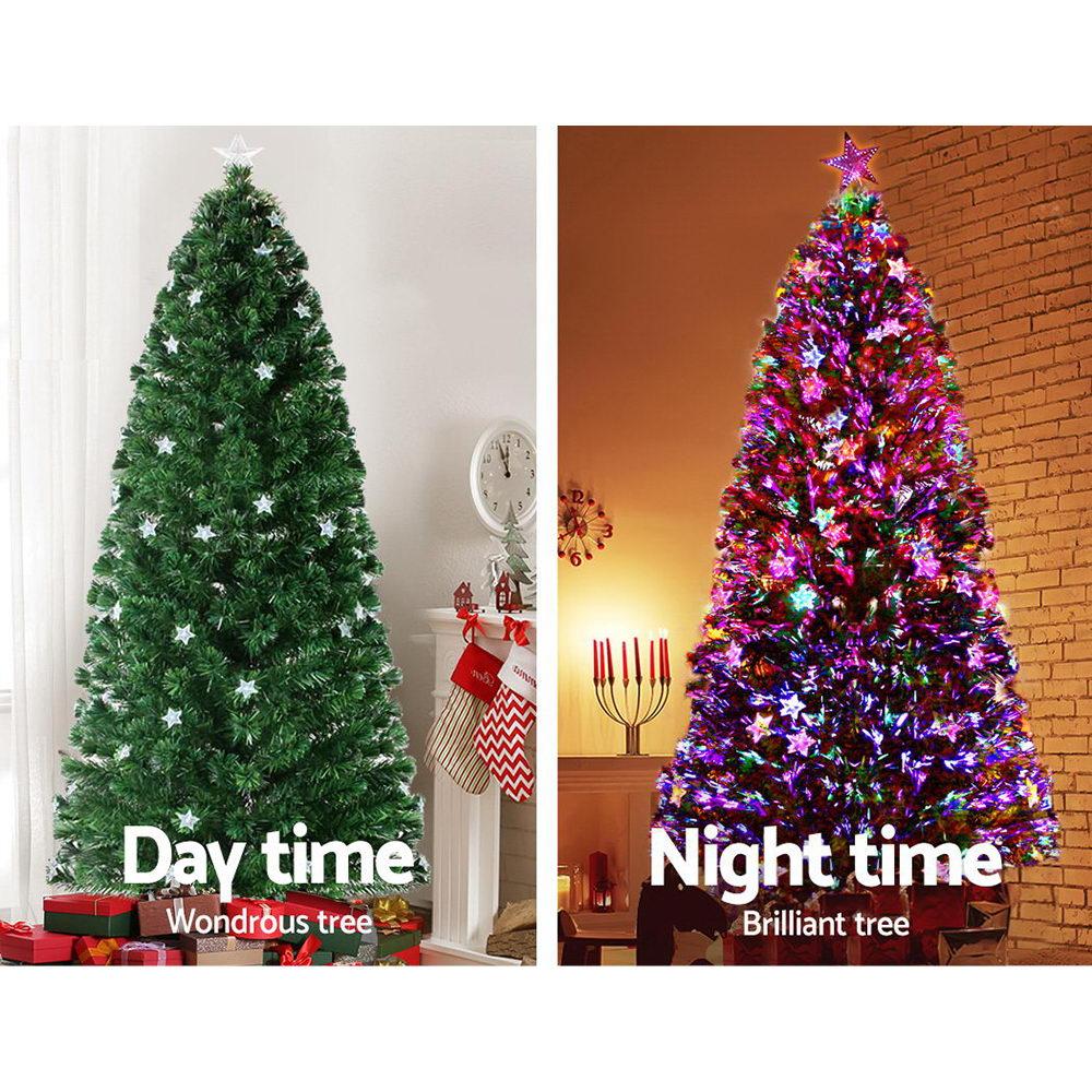 A split comparison showing a green artificial Christmas tree in two states: 'Day time' on the left, unlit and in a home setting, and 'Night time' on the right, illuminated with multicolored star-shaped lights against a brick wall background.