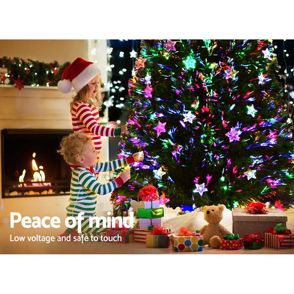 A festive Christmas scene with two children decorating a brightly lit Christmas tree near a fireplace. The tree is adorned with colorful star-shaped lights, and wrapped presents and a teddy bear sit at the base.