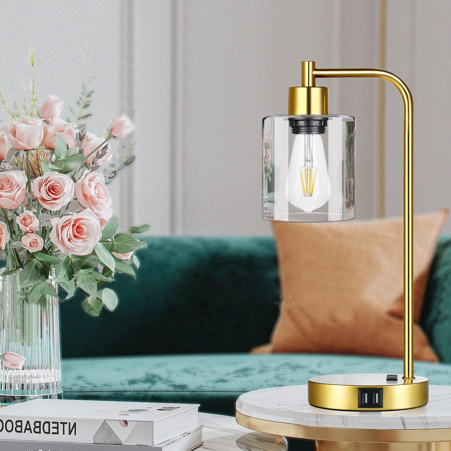Gold lamp with clear glass shade on a marble surface next to a vase of flowers and a green sofa.