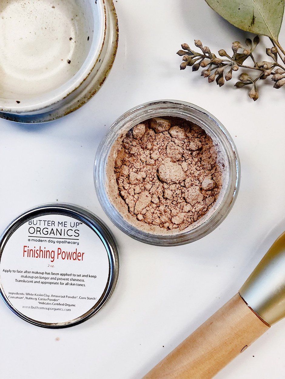 Butter Me Up Organics finishing powder jar with a brush and leaves on a white surface. deep hydrating body butter for winter dry skin and online store. Axis-Dynamics