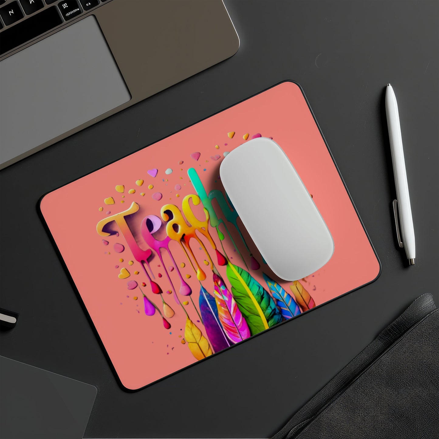 A rectangular desk mat with a coral pink background and a colorful dripping paint design spelling 'Teach' with a feather motif, shown on a dark desk with a white mouse on top, alongside a laptop corner, pen, and notebook. multi device wireless keyboard and mouse combo. Axis-Dynamics