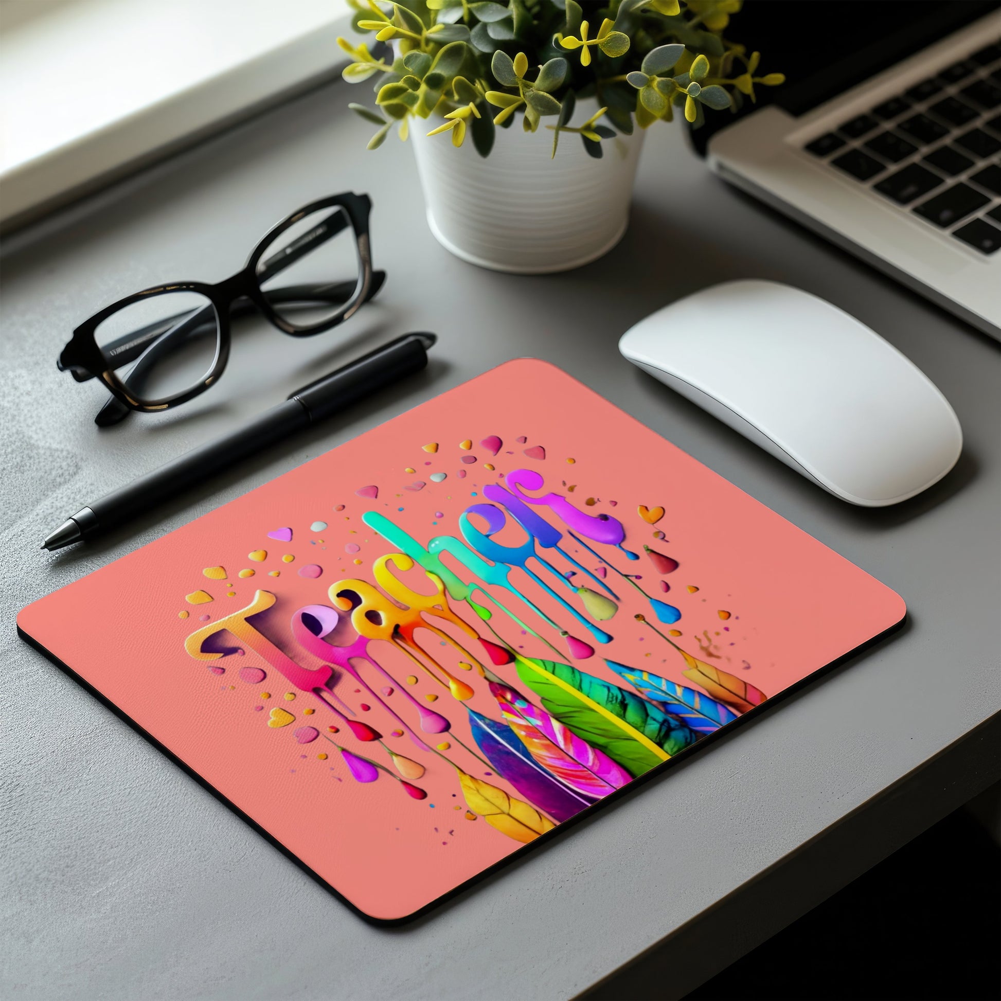 A rectangular desk mat with a coral pink background and a colorful dripping paint design spelling 'Lemon' in the center, shown on a gray desk with a white mouse, black pen, glasses, and a small potted plant. multi device wireless keyboard and mouse combo. Axis-Dynamics