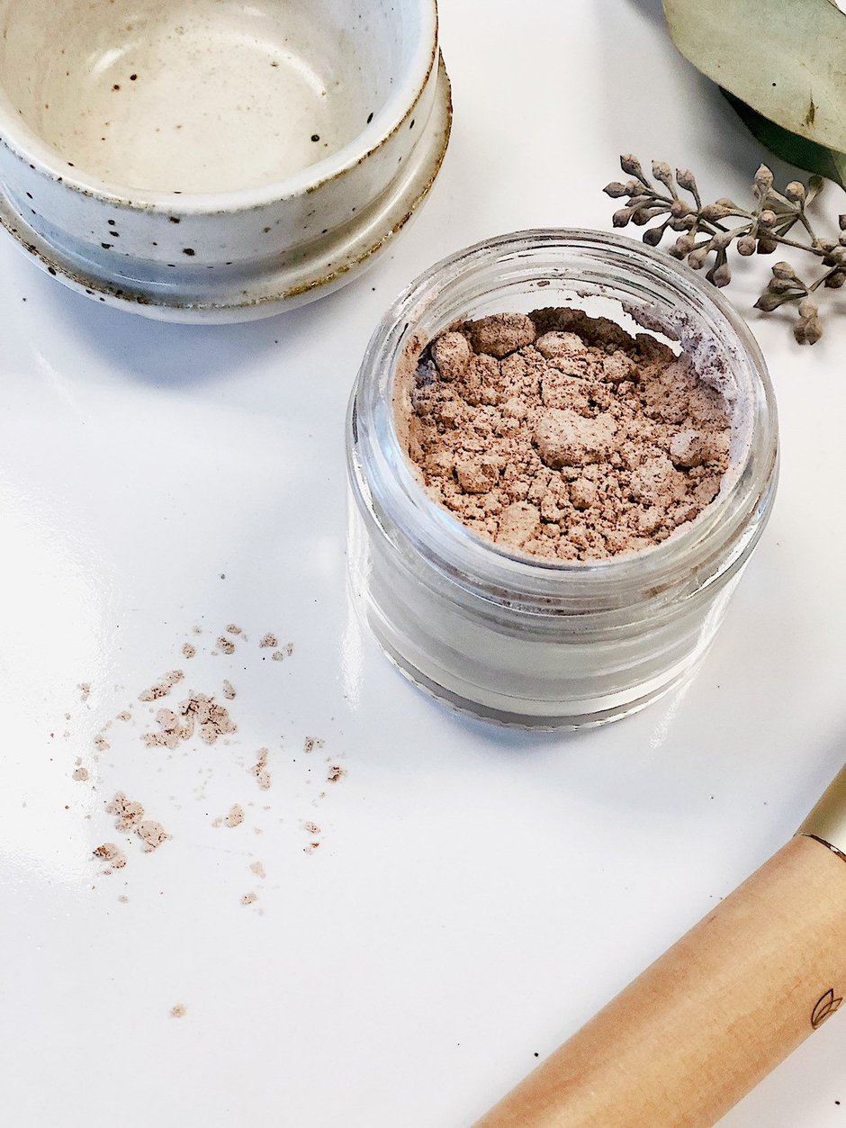 Jar of brown powder on a white surface with a small bowl and dried flowers. deep hydrating body butter for winter dry skin and online store. Axis-Dynamics
