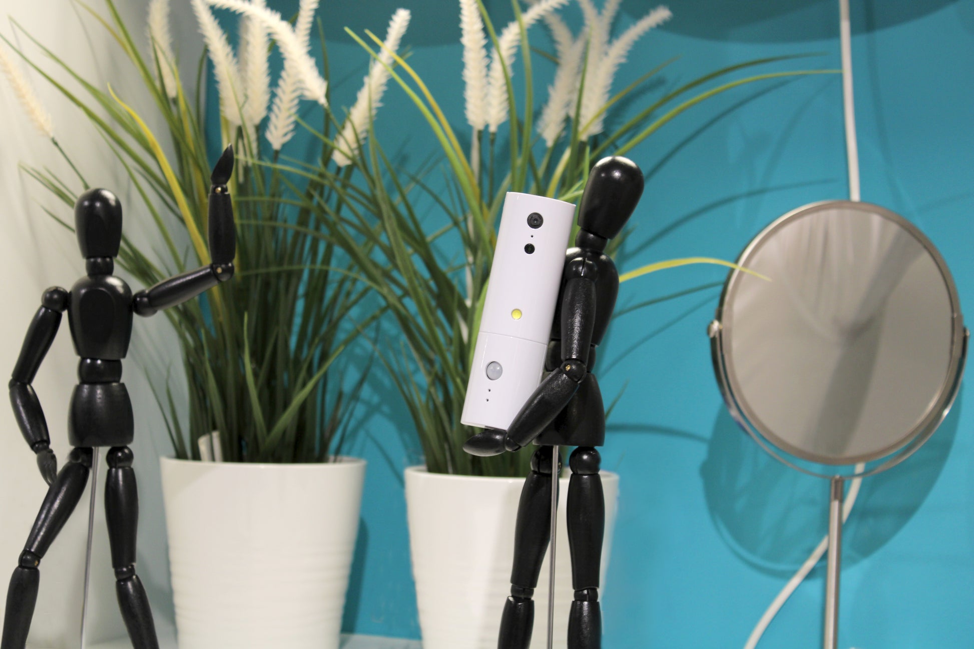 Two black mannequin figures in white pots with a plant, smartphone, and round mirror against a blue wall.