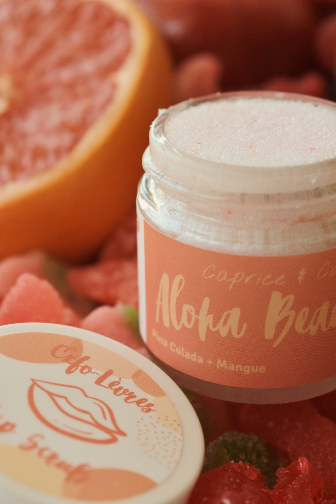 A close-up of a small clear jar filled with white scrub, with its lid placed in front. The jar has a peach label reading 'Aloha Beach' and 'Caprice + Co. Best online retail store. Axis-Dynamics