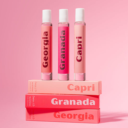 Three perfume spray bottles labeled Georgia, Granada, and Capri stand upright on stacked rectangular boxes in a pink studio setup. Best online retail store. Axis-Dynamics